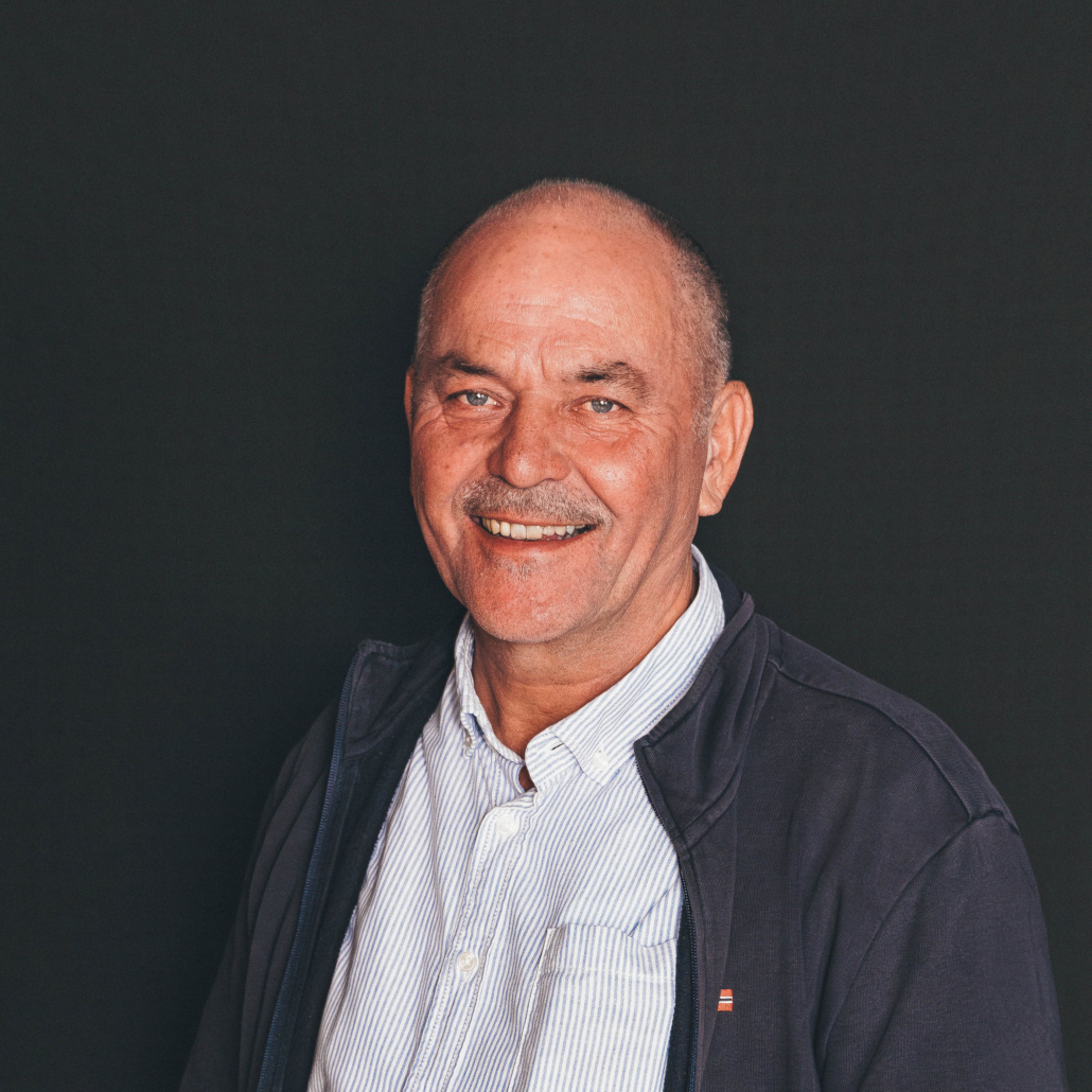 Ralf Einlehner Porträtfoto von Ralf Einlehner, der freundlich in die Kamera lächelt. Er trägt ein hellblau-weiß gestreiftes Hemd unter einer dunkelblauen Reißverschlussjacke vor einem einfarbigen, dunklen Hintergrund.