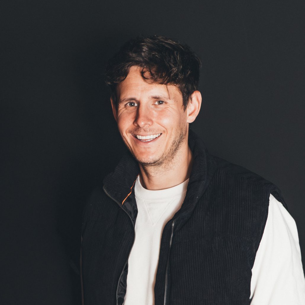 Tobi Krug "Porträtfoto von Tobi Krug, der lächelnd in die Kamera blickt. Er hat kurze, dunkle Haare und trägt ein weißes Sweatshirt unter einer schwarzen Kordweste. Der Hintergrund ist einfarbig und dunkel."