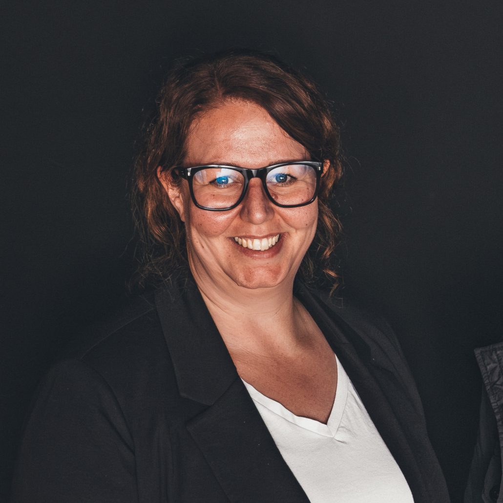 Mimi Hollmann Porträtfoto von Mimi Hollmann, die freundlich in die Kamera lächelt. Sie hat lockige, rötlich-braune Haare und trägt eine Brille mit dunklem Gestell, ein weißes T-Shirt und einen schwarzen Blazer. Der Hintergrund ist einfarbig und dunkel.