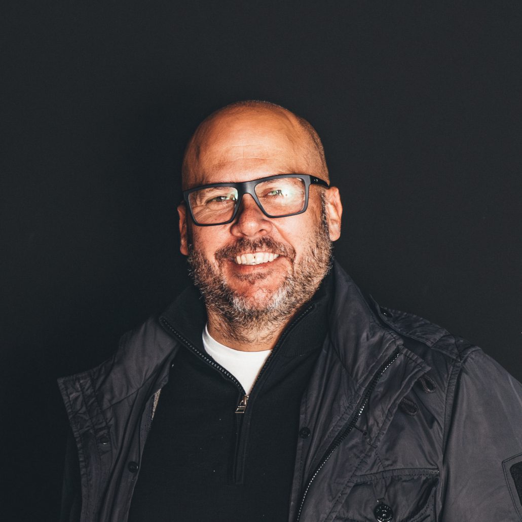 Heiko Didlapp Porträtfoto von Heiko Didlapp, einem Mann mit Brille und Bart, der lächelnd in die Kamera blickt. Er trägt eine dunkle Jacke über einem dunklen Pullover mit Reißverschluss. Der Hintergrund ist dunkel.