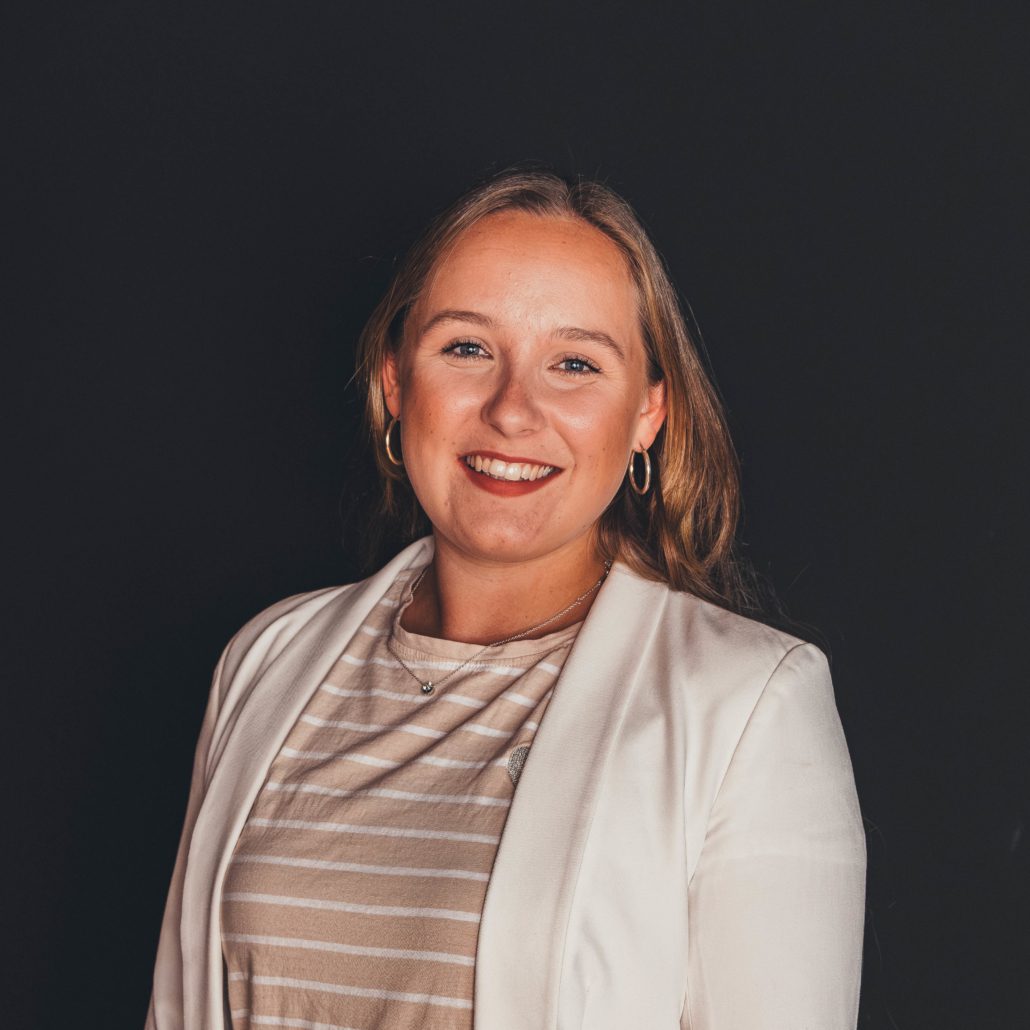 Julie Wagner Porträtfoto von Julie Wagner, einer jungen Frau, die lächelnd in die Kamera blickt. Sie trägt einen weißen Blazer über einem beige-weiß gestreiften Oberteil und Creolen-Ohrringe. Der Hintergrund ist dunkel.