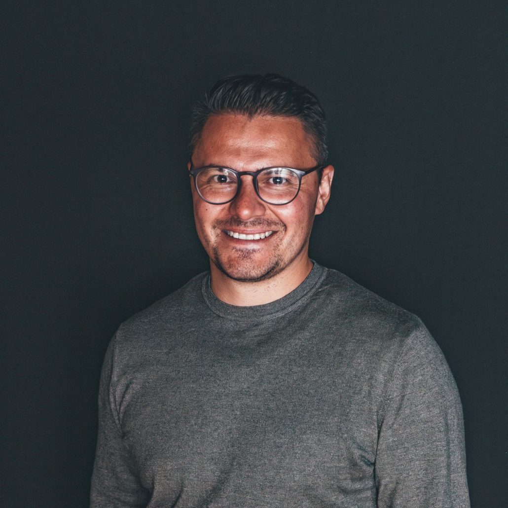 Viktor Meng Porträtfoto von Viktor Meng, einem Mann mit Brille, der lächelnd in die Kamera blickt. Er trägt einen grauen Pullover. Der Hintergrund ist dunkel.