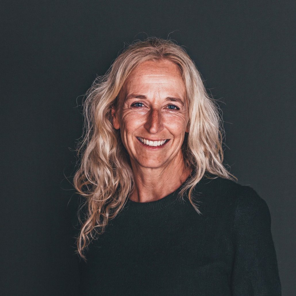 Rebekka Baumann Porträtfoto von Rebekka Baumann, einer Frau mit langen, welligen blonden Haaren, die lächelnd in die Kamera blickt. Sie trägt einen dunklen Pullover. Der Hintergrund ist dunkel.