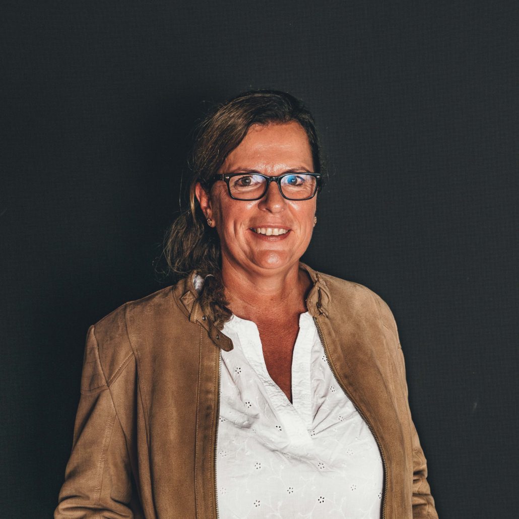 Christine Eidenmüller Porträtfoto von Christine Eidenmüller, einer Frau mit Brille, die lächelnd in die Kamera blickt. Sie trägt eine braune Jacke über einer weißen Bluse. Der Hintergrund ist dunkel.