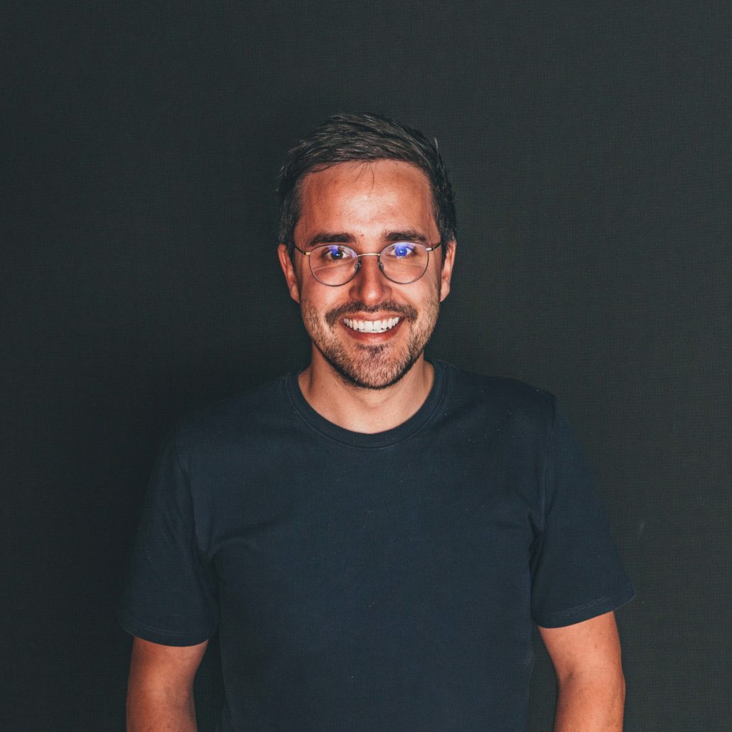 Sebastian Prinz Porträtfoto von Sebastian Prinz, einem Mann mit Brille, der lächelnd in die Kamera blickt. Er trägt ein dunkles T-Shirt. Der Hintergrund ist dunkel.