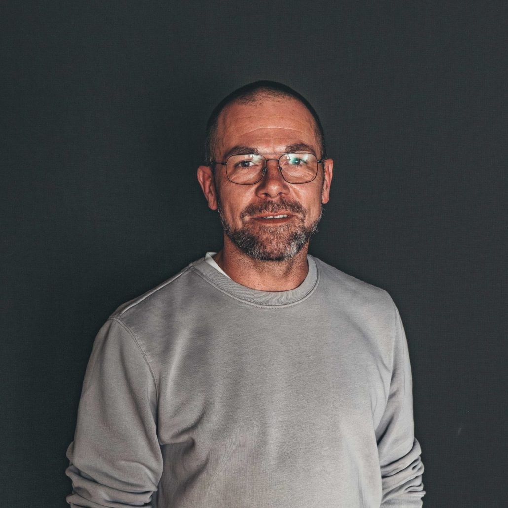 Marvin Brück Porträtfoto von Marvin Brück, einem Mann mit Brille und Bart, der in die Kamera blickt. Er trägt einen hellgrauen Pullover. Der Hintergrund ist dunkel.