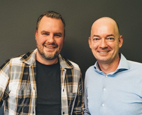 Foto-Flo-Christian Porträtfoto von Florian Kern (links) und Christian Muskat (rechts), die nebeneinander stehen und lächelnd in die Kamera blicken. Florian Kern trägt ein kariertes Hemd über einem schwarzen T-Shirt, Christian Muskat trägt ein hellblaues Hemd. Der Hintergrund ist dunkelgrau.